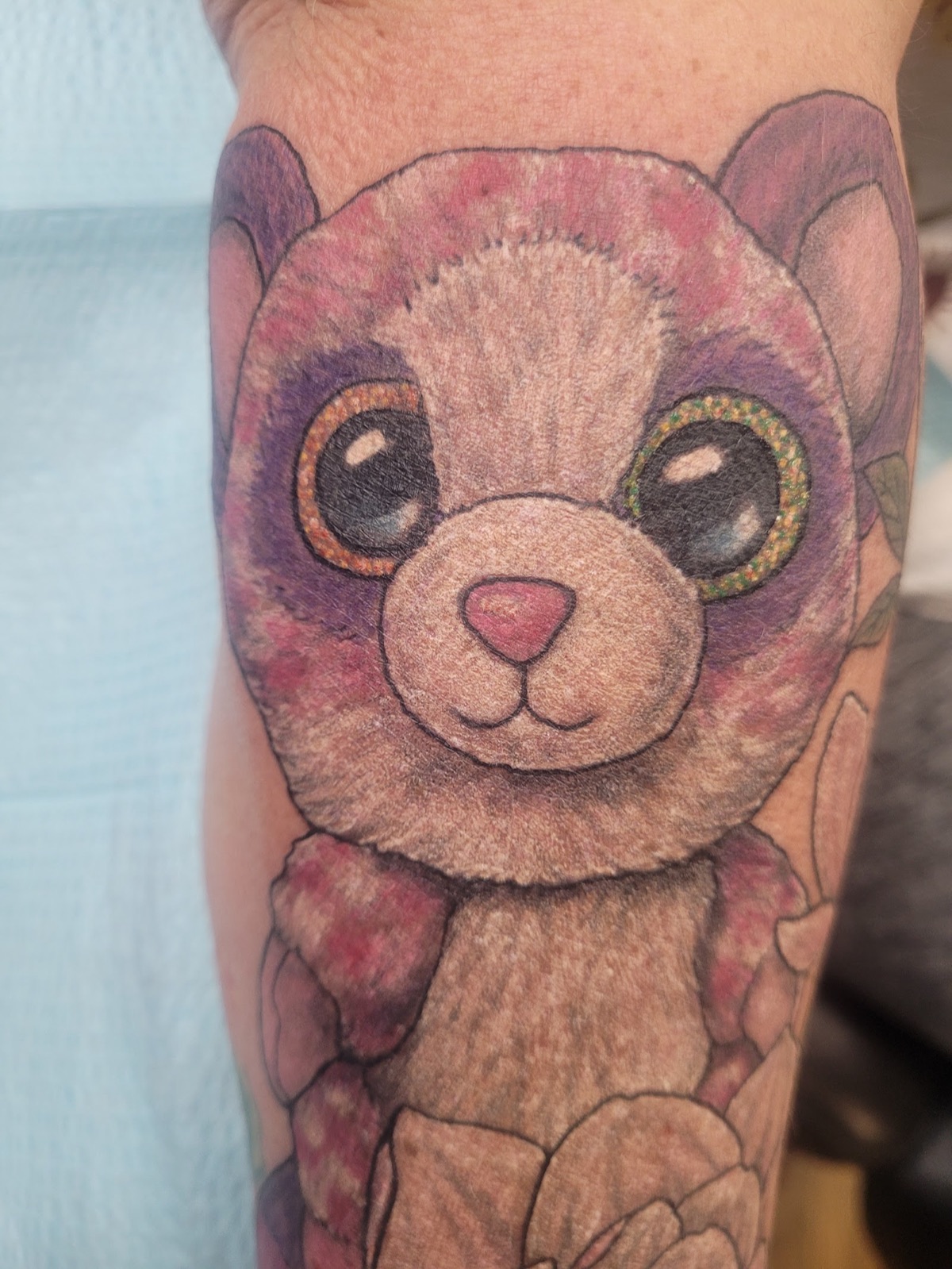 Color tattoo of a big-eyed plush teddy bear on a forearm.